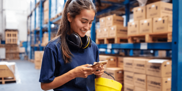 A woman working in a factory setting smiles while she uses her smartphone. 